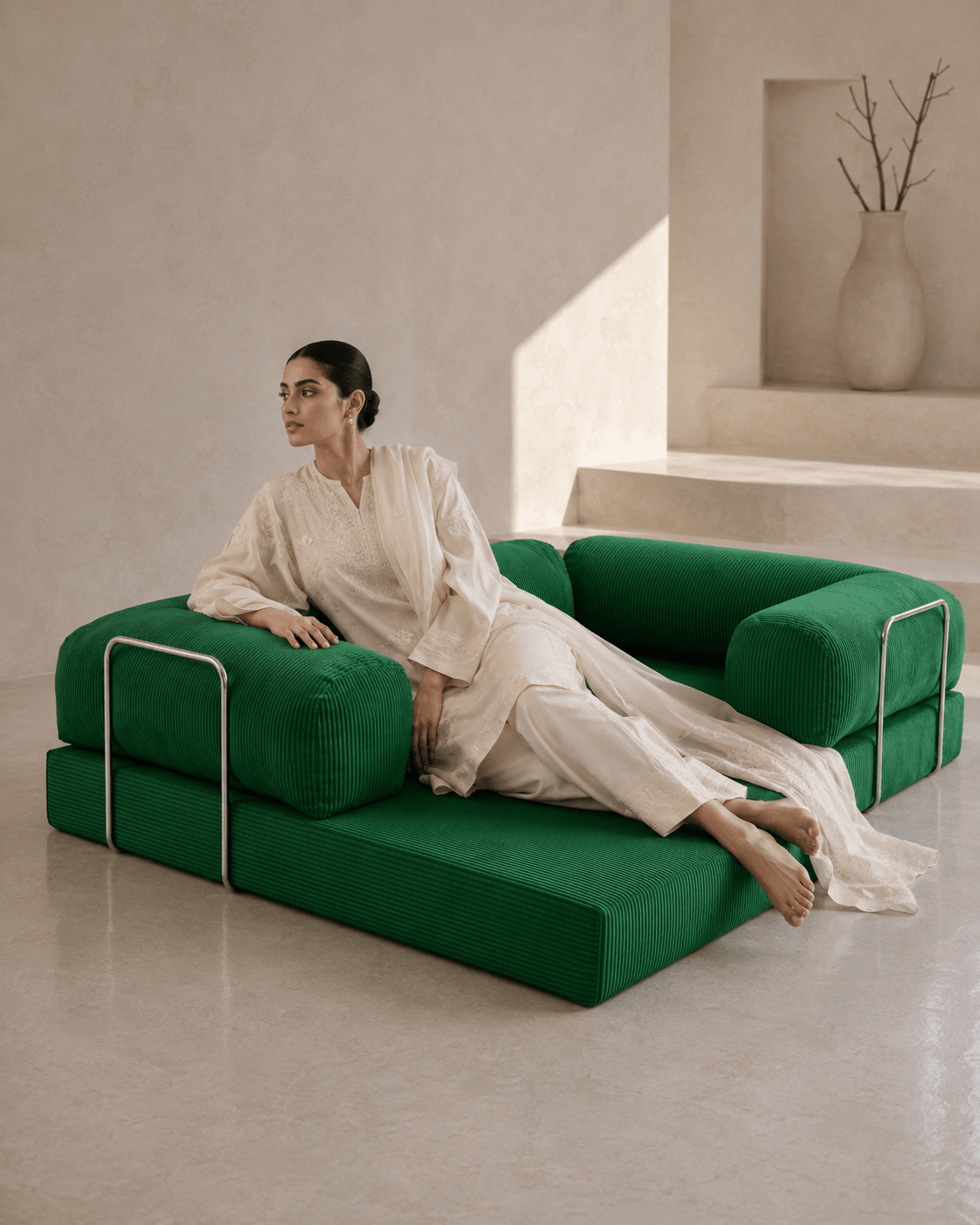 A woman in a white robe reclines on a modern modular green sofa with chrome metal frame accents, set in a minimalist beige interior room.